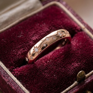 May include: A rose gold band ring featuring multiple star-shaped engravings, each set with a small, clear, round-cut diamond. The polished metal surface reflects light, highlighting the celestial-inspired pattern embedded along the outer circumference.