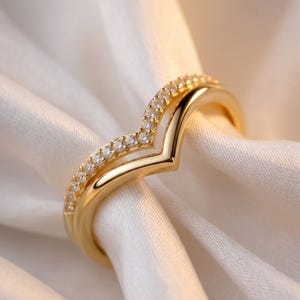 May include: A gold chevron-shaped ring featuring a polished band and a second parallel band encrusted with small, clear, round-cut stones. The ring has a V-shaped design that follows the contour of the finger, showcasing a bright metallic finish.