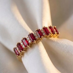 Op de afbeelding: Een gouden ring met een rij afwisselend geplaatste, baguette-geslepen rode robijnen en kleine ronde witte diamanten. De stenen zijn in een klauwzetting langs de bovenste helft van de ring geplaatst, wat een lijn van levendig rood en wit vormt.