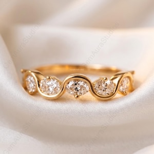 Puede incluir: Un anillo de oro con una serie de diamantes de talla ovalada dispuestos en un diseño ondulado. El anillo se muestra sobre una tela blanca y sedosa, resaltando su brillo y diseño.