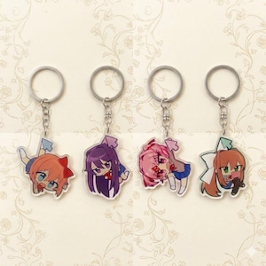 May include: Four anime-style character keychains. Each keychain features a different character with unique hair and clothing colors. The keychains are attached to silver-colored key rings and chains. The background is a cream color with a floral pattern.