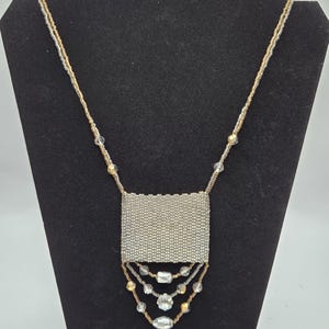 May include: A beaded necklace featuring a rectangular woven silver pendant suspended from a long chain of small metallic gold and silver beads. Three draped strands of clear faceted crystals and gold beads hang below the central pendant.