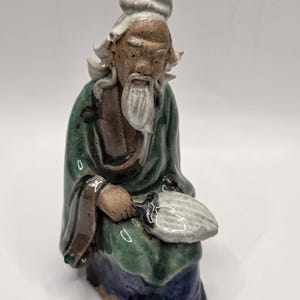 May include: A ceramic figurine of an elderly person with a long white beard and hair, wearing a green robe with blue accents. The figure is seated and holds a white fan in one hand, featuring a glossy glaze finish and detailed facial features.