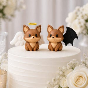 Könnte beinhalten: Zwei braune Fuchsfiguren auf einer weißen Torte. Der linke Fuchs hat weiße Engelsflügel und einen gelben Heiligenschein. Der rechte Fuchs hat schwarze Fledermausflügel. Beide Füchse haben dunkle Augen, eine helle Schnauze und eine matte Oberfläche.