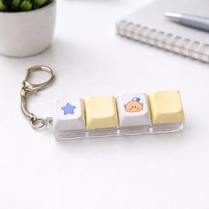 May include: A keyboard key-shaped keychain featuring four keys. Two keys are light yellow, one is white with a blue star, and another is white with a cartoon baby face. The keychain has a silver metal ring and chain.
