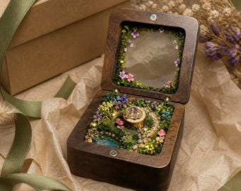 May include: A square dark wood ring box with a hinged lid featuring a glass panel framed by small pink and green floral accents. The interior contains a miniature garden landscape with moss, tiny colorful flowers, a stone path, and a ring.
