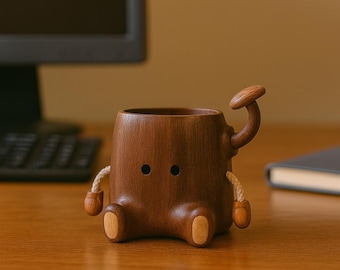 May include: A whimsical wooden mug with a unique design. The mug is brown with a mushroom-shaped handle and small, round eyes. It has wooden feet and arms with rope accents, creating a playful, cartoon-like appearance. The mug is sitting on a wooden surface.