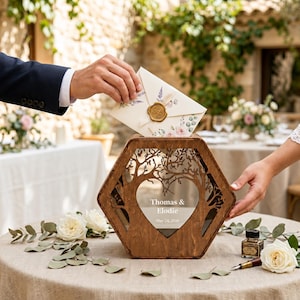 May include: A wooden wedding card box with a heart-shaped cutout and tree design. The box has the names "Thomas & Elodie" and the date "May 24, 2026" engraved on it. An envelope is being placed into the box.