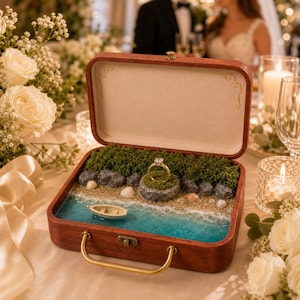 May include: A wooden ring box featuring a miniature beach diorama with blue resin water, a small boat, sand, rocks, and green moss. A gold ring with a clear stone sits atop a mossy rock in the center. The box has a metal latch and handle.