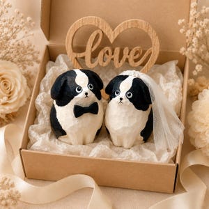 May include: Two carved wooden dog figurines in a cardboard gift box. One dog wears a black bow tie, and the other wears a white veil with small pearls. A wooden heart-shaped cutout with the word love is positioned behind the two figures.