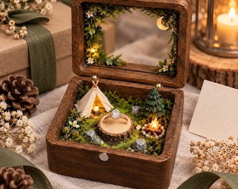 May include: A dark wood ring box featuring a miniature camping scene with a tent, a campfire, a pine tree, and moss. A gold ring with a clear stone sits on a wooden stump in the center. The lid interior is decorated with moss, flowers, and a crescent moon.