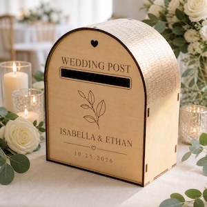 May include: A light-colored wooden wedding post box with an arched top, featuring a horizontal slot, a small heart icon, a leaf illustration, and the engraved text "WEDDING POST", "ISABELLA & ETHAN", and the date "10.25.2026" on the front panel.