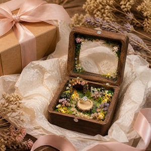 Pode incluir: Uma caixa de anel quadrada de madeira com tampa de vidro articulada, apresentando um jardim em miniatura no interior com musgo, pequenas flores, uma cerca branca e um pedestal central de madeira com um anel dourado de pedra clara.