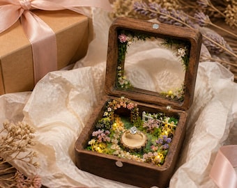 May include: A square wooden ring box with a hinged glass lid, featuring an interior miniature garden scene with moss, tiny flowers, a small white fence, and a central wooden pedestal holding a gold ring with a clear stone, illuminated by small lights.
