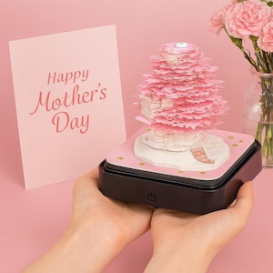May include: A pink paper art sculpture with a layered tree design, illuminated on a black base. A pink card with "Happy Mother's Day" is visible. A vase of pink flowers is in the background.