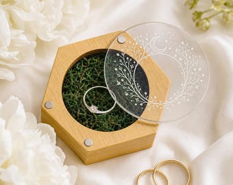May include: A hexagonal wooden ring box with a circular interior filled with green moss. A silver ring with a clear stone sits inside. The lid is a clear acrylic disc featuring a white etched crescent moon and floral wreath design.