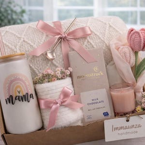May include: A gift basket with a white tumbler featuring a rainbow design and the word "Mama". The basket includes a pink bow, a small spoon, a candle, a chocolate bar, and a crochet tulip. The basket is filled with other decorative items.