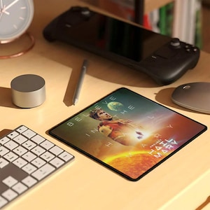 May include: A black mousepad with the text "BELIEVE IN THE HAIL MARY" and an image of a person in a red and yellow uniform. A black handheld gaming device, a silver mouse, and a keyboard are also on the desk.