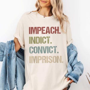 May include: Cream-colored t-shirt with the words "IMPEACH. INDICT. CONVICT. IMPRISON." in a distressed, vintage-style font. The shirt is paired with a denim jacket.