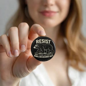 May include: A black button with the word "RESIST" in white block letters. Below the word is a detailed illustration of a bear. The text "WHEN TYRANNY BECOMES LAW REBELLION BECOMES DUTY" is printed below the bear.