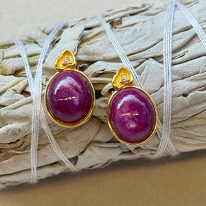 May include: Gold-coloured earrings with oval, deep magenta gemstones. Each earring has a small, yellow flower-shaped accent with a tiny diamond. The earrings are displayed on a bundle of dried sage.