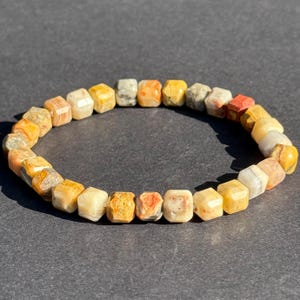 Può includere: Un braccialetto di perline cubiche multicolori, con sfumature di giallo, arancione, rosso e grigio. Il braccialetto è esposto su una superficie grigio scuro. Le perline misurano circa 1 cm.