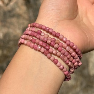 May include: A stack of six pink gemstone bracelets. Each bracelet is made of small, faceted, square-shaped beads. The bracelets are wrapped around a wrist, with the skin tone of the wrist visible.