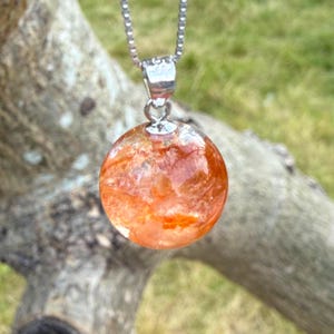 Op de afbeelding: Een ketting met een zilveren ketting en een ronde, oranje hanger. De hanger is een heldere bol met een oranje, getextureerd interieur. De ketting wordt getoond tegen een vage achtergrond van groen gras en een tak.