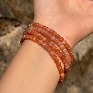 May include: A stack of four bracelets made of small, faceted, orange-red beads. The bracelets are worn on a wrist, with the beads catching the light and displaying a range of warm tones.