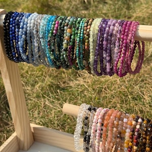 Könnte beinhalten: Eine Präsentation von Armbändern mit Perlen in verschiedenen Farben, darunter Schwarz, Blau, Grün, Lila und Rosa. Die Armbänder sind auf einem Holzdübel aufgefädelt, wodurch die Vielfalt der Edelsteine und Perlen zur Geltung kommt. Die Armbänder werden auf einem Holzständer ausgestellt.