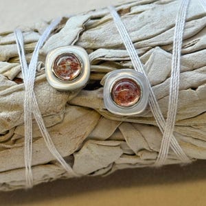 May include: Two square stud earrings with a central, reddish-brown stone set in a silver-coloured bezel. The earrings are displayed on a bundle of dried sage, with white string wrapped around it.