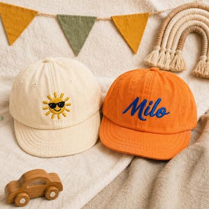 May include: Two baseball caps, one cream-coloured with an embroidered yellow sun wearing black sunglasses, and one orange cap with the name Milo embroidered in blue script. The caps feature a structured crown, a curved brim, and top stitching.