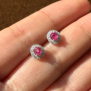 Peut inclure: Deux boucles d'oreilles à clous ornées de pierres rouges ovales vives, entourées de petites pierres claires. Les boucles d'oreilles sont serties dans un métal argenté, créant un design classique et élégant.