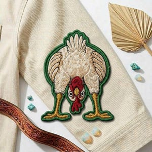 May include: Embroidered patch featuring a chicken in a comical pose, with its head down and rear end up. The patch has a green border and is sewn onto a cream-colored fabric. The chicken has a red comb and yellow legs. A brown leather belt and decorative items are in the background.