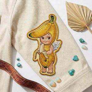 May include: Embroidered patch featuring a whimsical banana-themed cherub with wings, holding a peeled banana. The patch is primarily yellow with green and brown accents, and is attached to a cream-colored jacket.