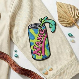 May include: Embroidered patch of a soda can with the word "NAME" in pink, blue, and green. The patch is on a cream-colored jacket sleeve. Decorative elements include a brown leather belt, a dried leaf, and small stones.