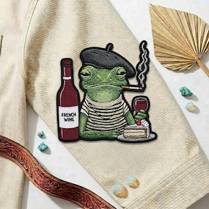 May include: Embroidered patch featuring a green frog wearing a beret and striped shirt, smoking a cigarette, and holding a glass of wine. A bottle of French wine and a slice of cake are also present. The patch is on a beige jacket.