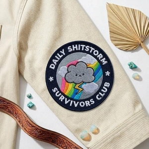 May include: A round patch with the text "DAILY SHITSTORM SURVIVORS CLUB" encircling a cartoon cloud, rainbow, and lightning bolt design. The patch is sewn onto a cream-colored jacket sleeve. Decorative elements include a leather belt and small gemstones.
