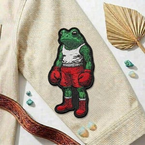 May include: Embroidered patch featuring a green frog in a boxer's outfit. The frog wears a white tank top, red boxing gloves, and red shorts and boots. The patch is on a beige fabric.