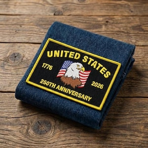May include: A folded denim item with a rectangular patch featuring the text "UNITED STATES" and "250TH ANNIVERSARY". The patch includes an eagle, the American flag, and the dates 1776 and 2026. The patch has a yellow border.