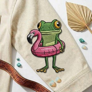 May include: Embroidered patch featuring a green frog wearing a pink flamingo floatie. The frog has large yellow eyes and is set against a beige fabric background. A brown leather belt and decorative leaf are also visible.