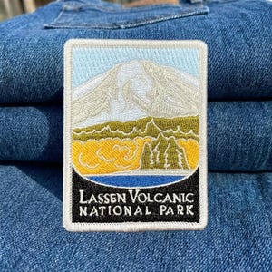 May include: Embroidered patch featuring a mountain landscape with a light blue sky, a white mountain, and a yellow and green foreground. The patch has the text "Lassen Volcanic National Park" in white letters on a black background.