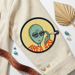 May include: Embroidered patch featuring a green alien smoking a joint, with the text "AREA 420" in orange. The patch is circular with a yellow background and a navy blue border, sewn onto a beige jacket.