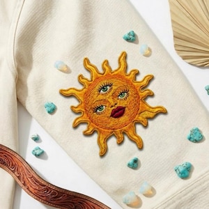 May include: Cream-colored jacket with an embroidered sun design featuring three eyes and red lips. The jacket is adorned with turquoise and opal stones, and a brown leather belt is visible.