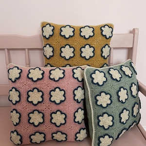 Könnte beinhalten: Drei gehäkelte Kissen in Senfgelb, Rosa und Salbeigrün. Jedes Kissen hat ein sich wiederholendes Muster aus weißen Blumenformen, die mit Marineblau umrandet sind. Die Kissen sind quadratisch und scheinen handgefertigt zu sein.
