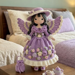 Könnte beinhalten: Eine handgefertigte Häkelpuppe mit dunklem Haar, einem lavendelfarbenen Hut mit breiter Krempe und einem violetten Kleid, das mit kleinen Stoffrosen verziert ist. Die Puppe hat große Flügel, einen passenden Beutel und ein Miniatur-Teeservice.