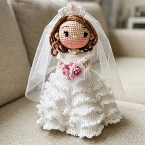 Könnte beinhalten: Eine handgefertigte Häkelpuppe in einem weißen, mehrlagigen Kleid mit passendem Schleier und Diadem. Die Puppe hat braunes Haar, eine Perlenkette und hält einen kleinen Strauß aus rosa gehäkelten Blumen. Das weiße Kleid ist mit Rüschen verziert.