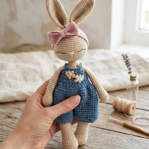 Könnte beinhalten: Gehäkelte Hasenpuppe mit beigem Körper, langen Ohren und rosa Schleife. Der Hase trägt einen blauen Overall mit Blumenapplikationen. Die Puppe wird in einer Hand auf einer Holzoberfläche gehalten.