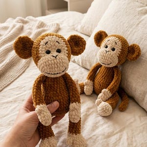 Könnte beinhalten: Zwei handgefertigte gehäkelte Affen-Plüschtiere. Einer wird von einer Hand gehalten, der andere sitzt auf einem Bett. Die Affen haben braune Körper, beige Gesichter, Ohren, Hände und Füße. Sie haben schwarze Augen und lächelnde Gesichter. Der Hintergrund ist ein neutral getöntes Schlafzimmer.