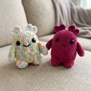 May include: Two small, handmade crochet plush toys with rounded bodies, short arms, and two-lobed head protrusions. One plush features a multicolored pastel yarn pattern of pink, yellow, and blue, while the other is a solid, deep magenta color.